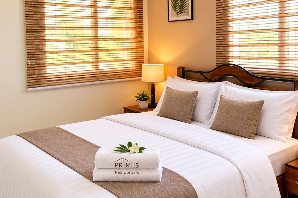 Un lit bien fait, recouvert d’une couverture blanche et marron, avec un oreiller et une serviette pliée ornée d’un logo.