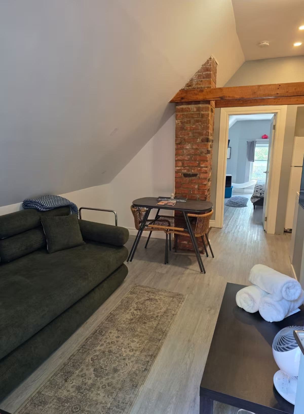 Un espace de vie de style loft avec un plafond haut, un mur d'accent en briques, un canapé vert, une petite table à manger avec des chaises en osier et une vue sur la pièce adjacente.