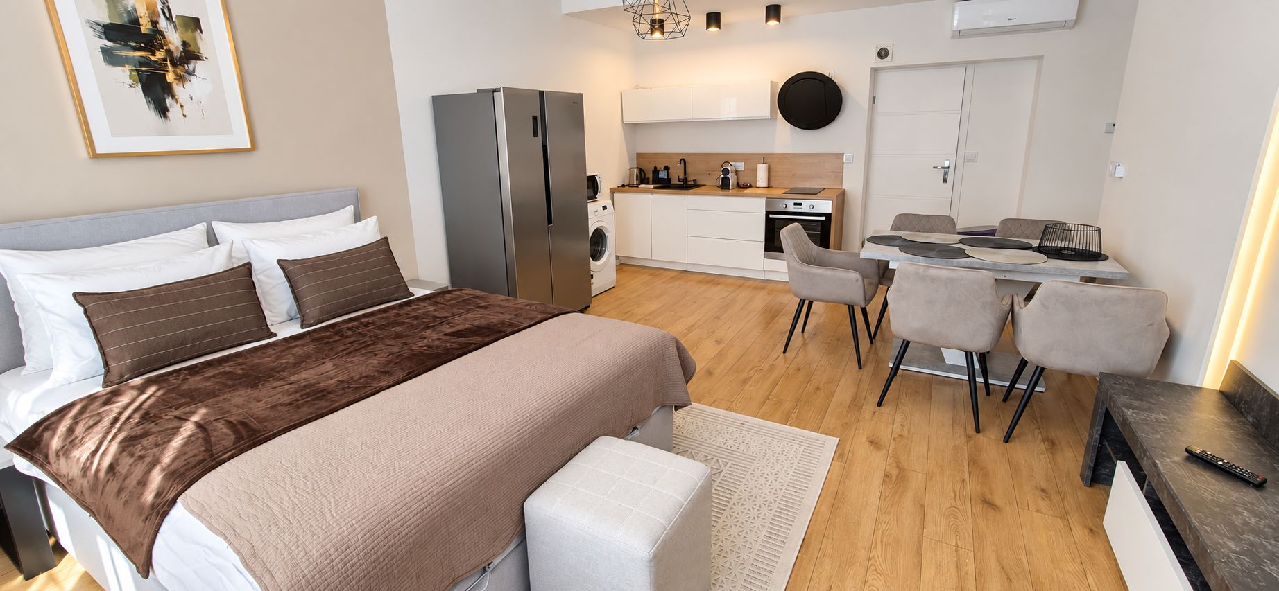 Un dormitorio moderno con una cama grande, una sala de estar separada con una mesa y sillas, lavadora incorporada y kitchenette.