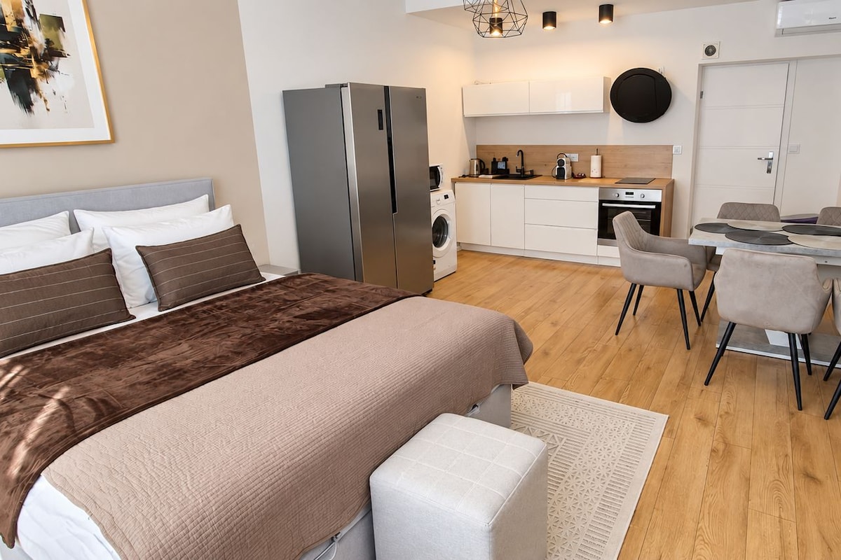 A modern bedroom with a large bed, a separate sitting area with a table and chairs, a built-in washing machine, and a kitchenette.