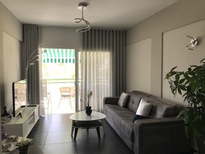 A modern living room with a sofa, a coffee table, and a television. There is a sliding glass door leading to an outdoor area. - Ocean Breeze Apartment on El Duque Beach (Costa Adeje)