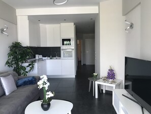 A modern living room with a sofa, a coffee table with flowers, a TV, and a kitchen area with white cabinets and appliances. - Ocean Breeze Apartment on El Duque Beach (Costa Adeje)