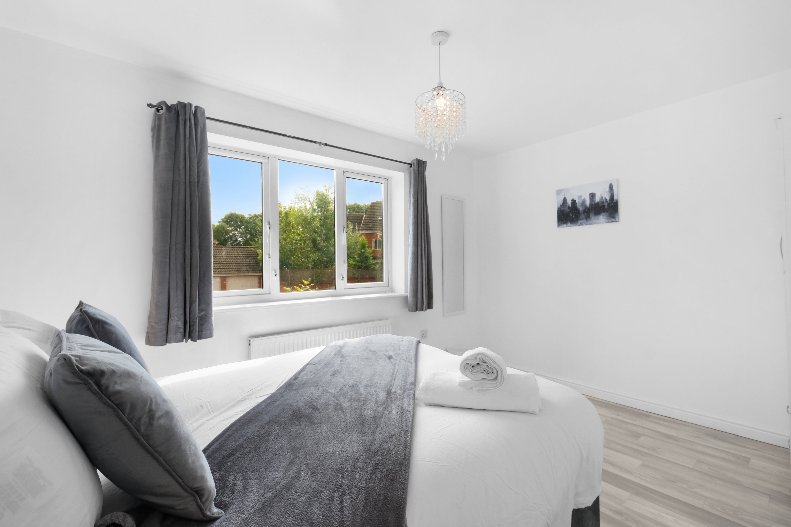 A bedroom with a bed, a window, a radiator, a chandelier, and a cityscape painting.