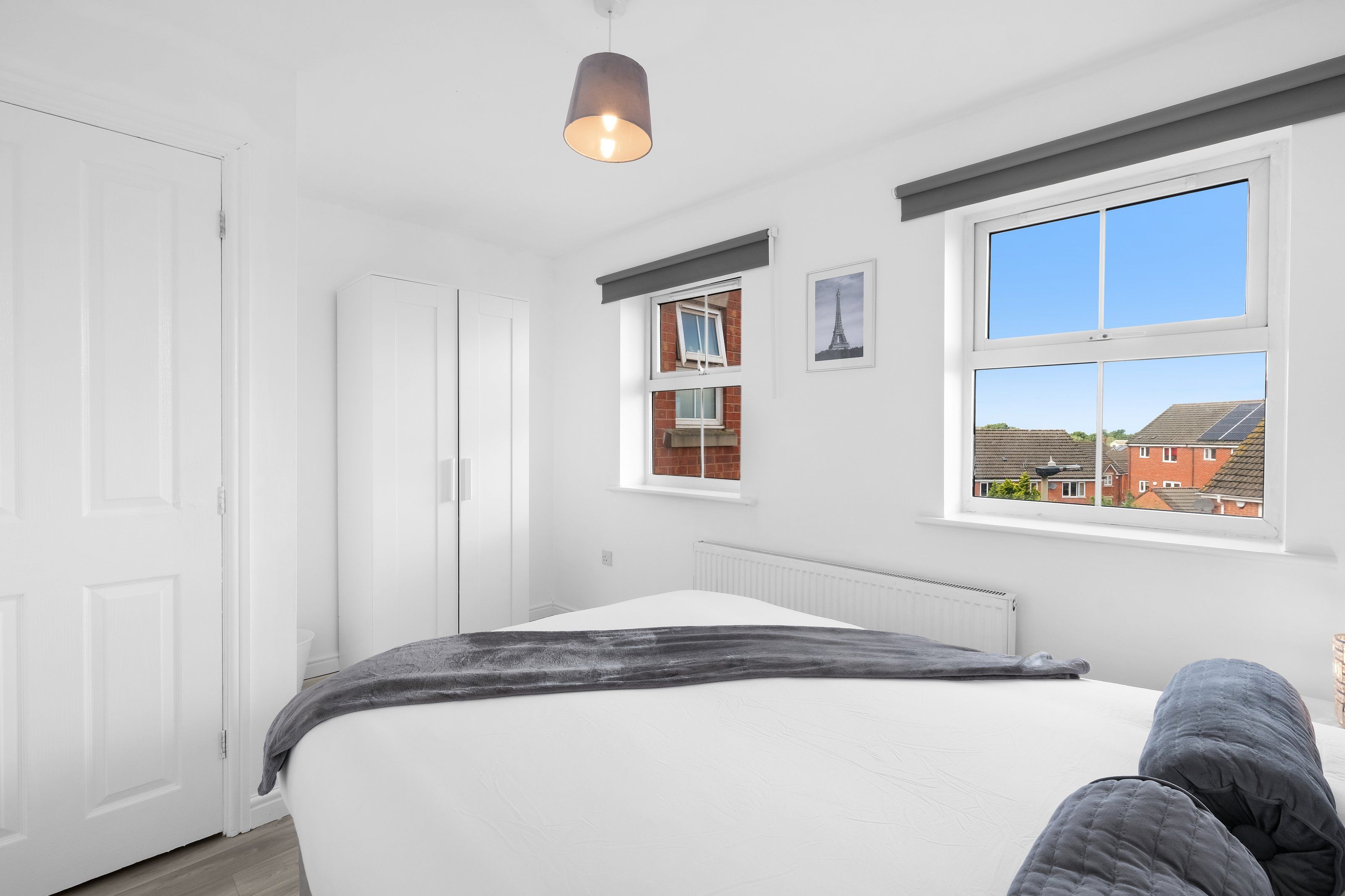 Ein Schlafzimmer mit einem Bett, einem Fenster mit Blick auf Gebäude und einer Deckenleuchte.