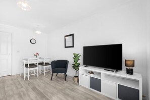 A modern living room with a flat-screen TV, a dining table with chairs, a potted plant, and a lamp. - 4-Bed Townhouse in Hamilton with Parking (Leicester)