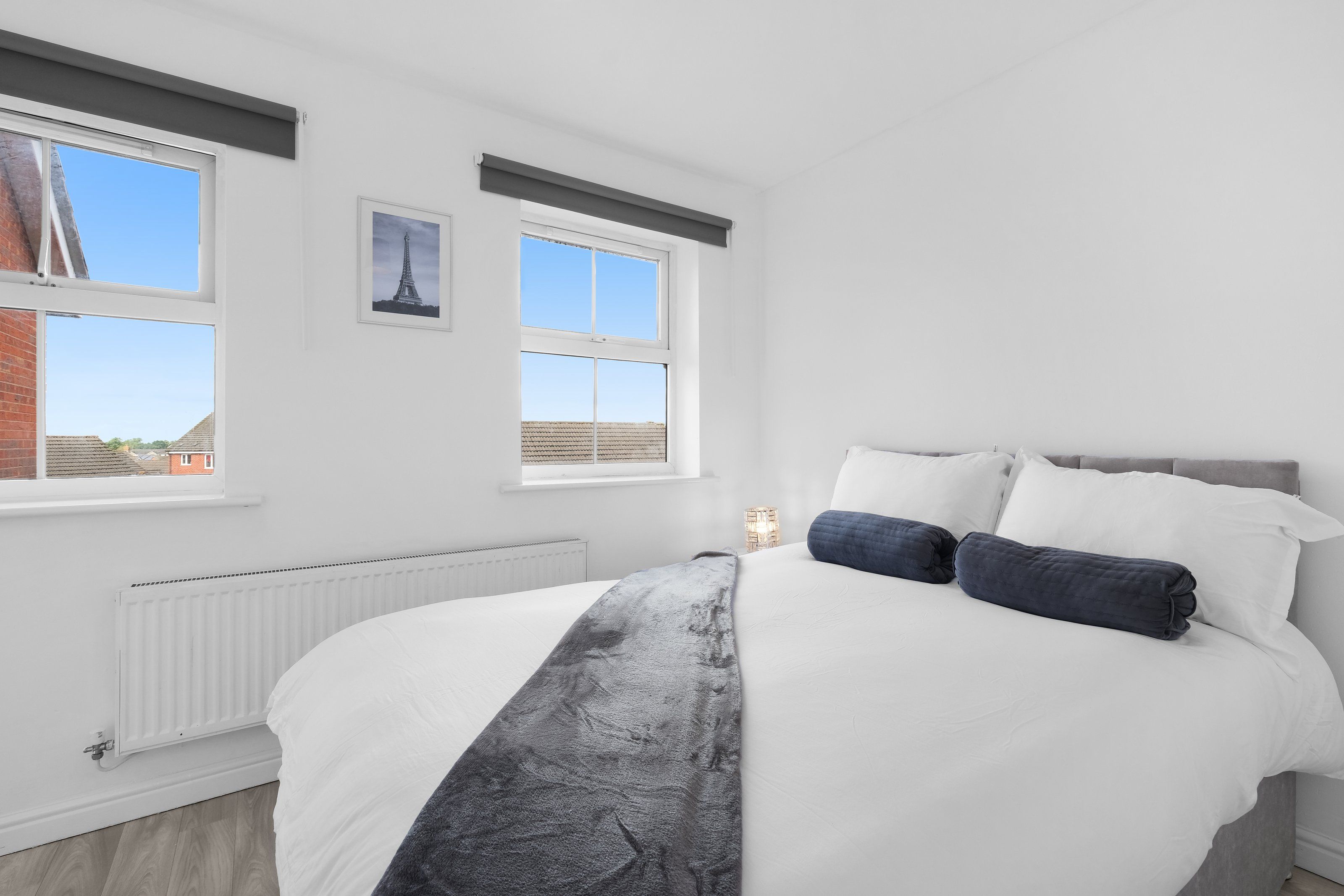 A bedroom with a large bed, two windows, a radiator, and a picture of the Eiffel Tower.