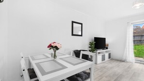 Ein modernes, minimalistisches Wohnzimmer mit einem weißen Tisch, schwarzen Stühlen, einem Flachbildfernseher und einem großen Fenster.