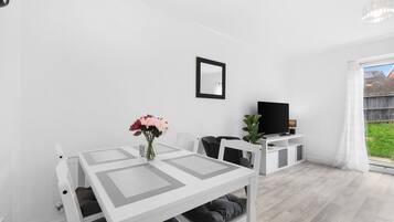 Una sala de estar moderna y minimalista con una mesa blanca, sillas negras, un televisor de pantalla plana y un ventanal grande.