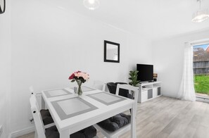 A modern, minimalist living room with a white table, black chairs, a flat-screen TV, and a large window. - 4-Bed Townhouse in Hamilton with Parking (Leicester)