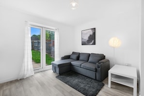 A modern living room with a grey sectional sofa, a white side table, a black rug, a framed artwork, and a floor lamp. - 4-Bed Townhouse in Hamilton with Parking (Leicester)