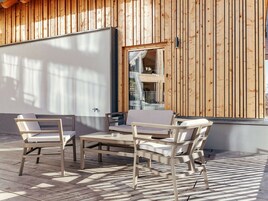 Un área de estar al aire libre con mesa y sillas, paredes de paneles de madera y vista a un edificio con balcón.