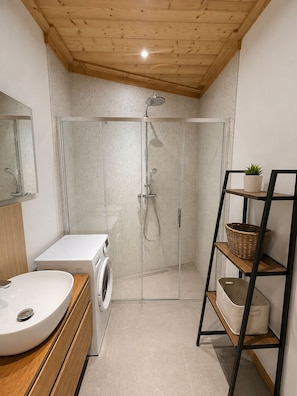 A modern bathroom with a wooden ceiling, a glass-enclosed shower, a white sink, a washing machine, and a shelf with baskets. - New Condo / Sleeps 4 / Right on the slopes - Les Menuires (Les Belleville)