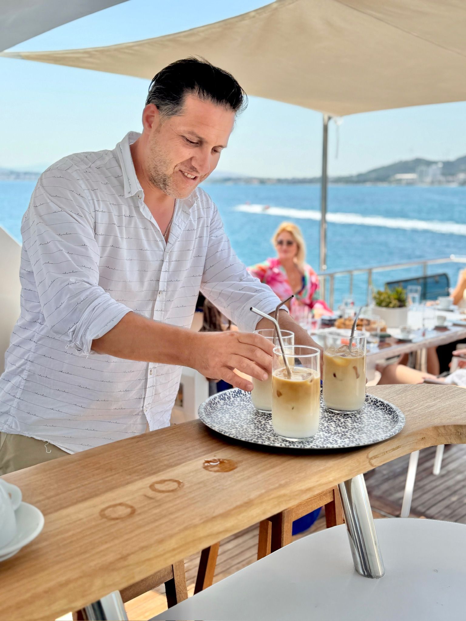 Eine Person serviert Eisgetränke an einem Holztisch mit Blick auf das Meer im Hintergrund.
