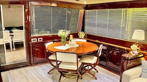 A spacious dining area with a round table and chairs, a small side table with a lamp, and a view through large windows. - 7 cabins 27M Benetti luxury yacht Velora (İzmir)