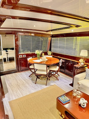A spacious dining area with a round table and chairs, a small side table with a lamp, and a view through large windows. - 7 cabins 27M Benetti luxury yacht Velora (İzmir)