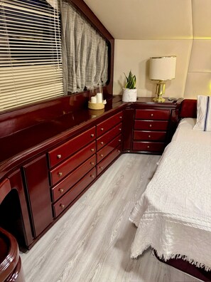A bedroom with a wooden dresser, a bedside table with a lamp, a bed with a white bedspread, and a window with blinds.
