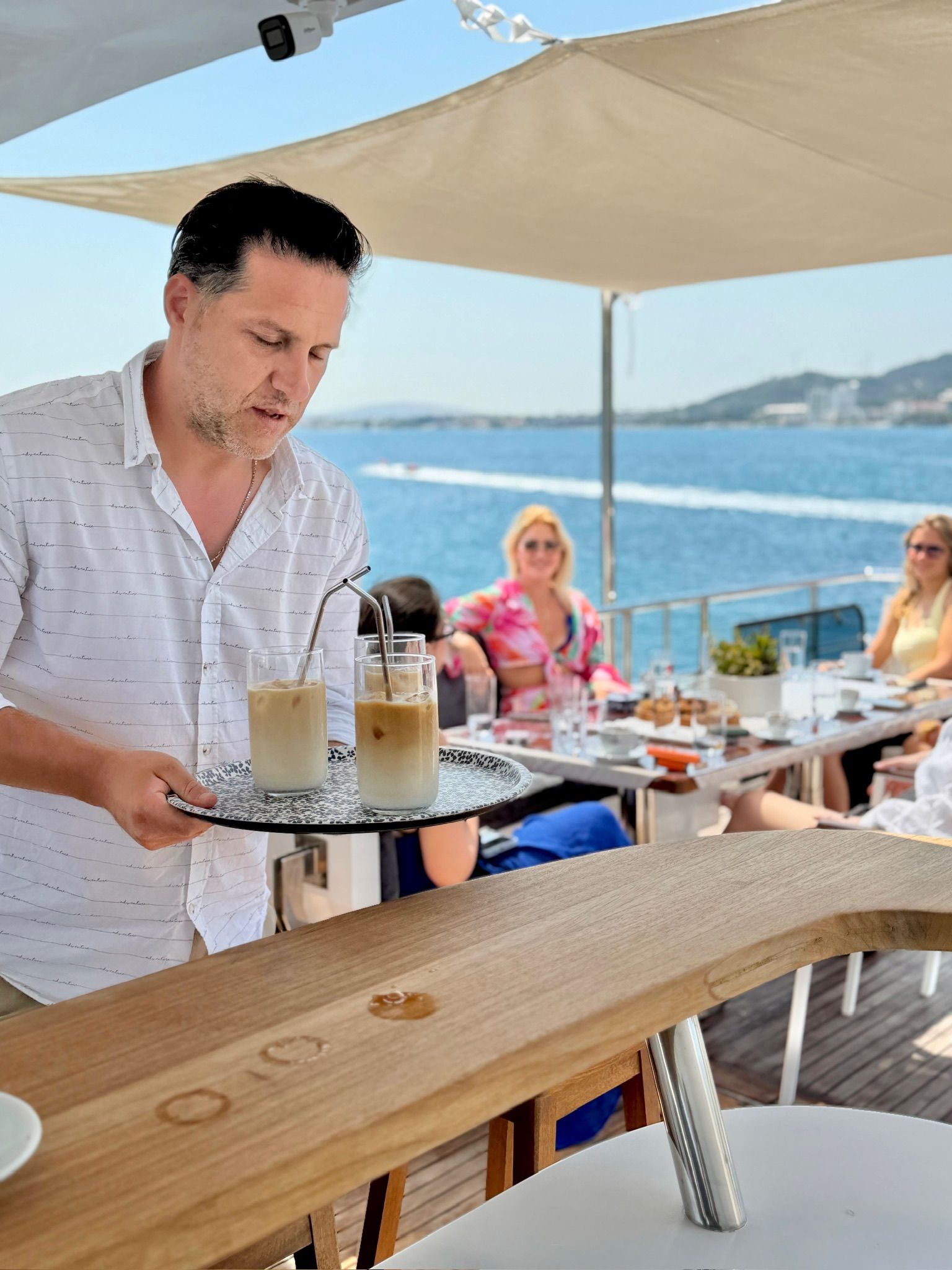 Ein Kellner serviert eisgekühlte Getränke an einem Holztisch mit Blick auf das Meer.