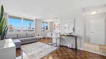 A modern living room with a sofa, dining table, chairs, a coffee table, and a large window with a view of buildings.