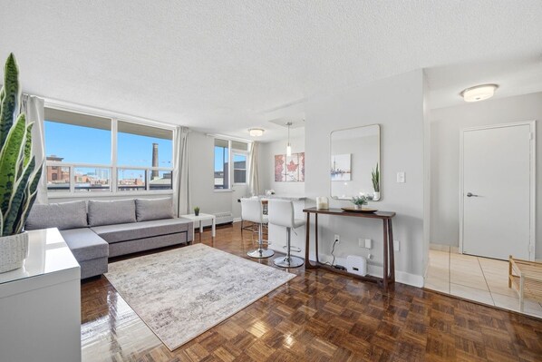 A modern living room with a sofa, dining table, chairs, a coffee table, and a large window with a view of buildings.