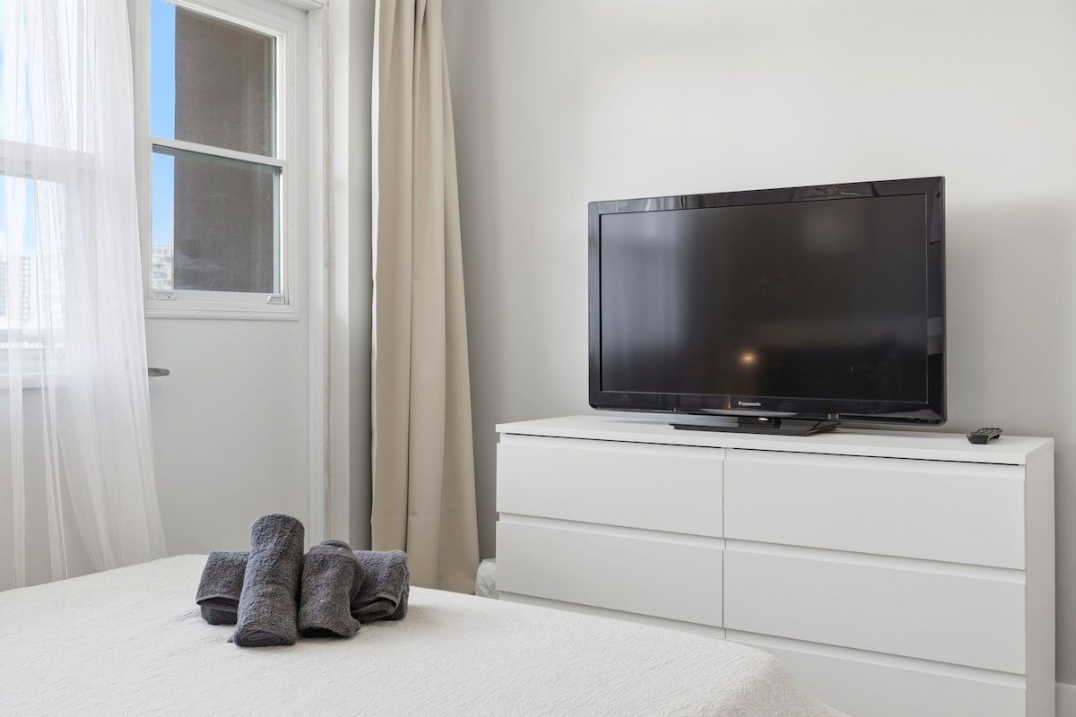 A bedroom with a bed, a television on a white dresser, and a window with curtains.