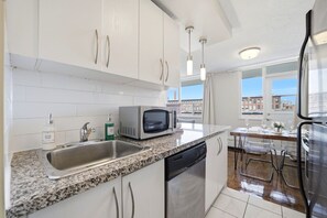 A modern kitchen with white cabinets, a granite countertop, a microwave, a dishwasher, and a view of buildings through large windows.