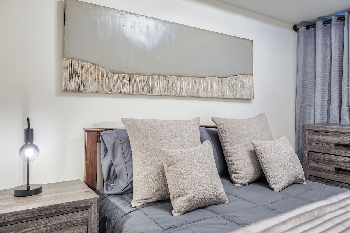 Quarto moderno com um sofá-cama cinza, mesa de cabeceira com abajur e um grande quadro abstrato na parede.