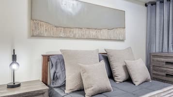 Quarto moderno com um sofá-cama cinza, mesa de cabeceira com abajur e um grande quadro abstrato na parede.