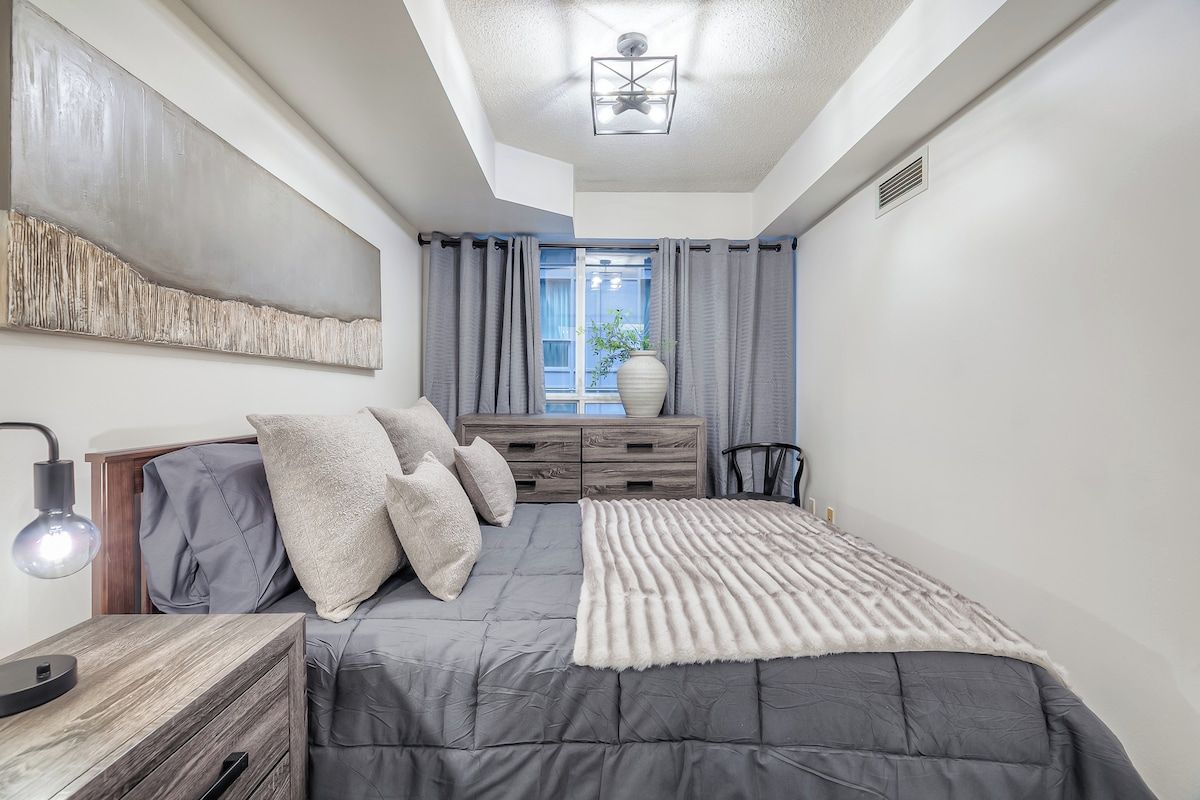 A bedroom with a bed, bedside table, lamp, dresser, and a window with curtains.