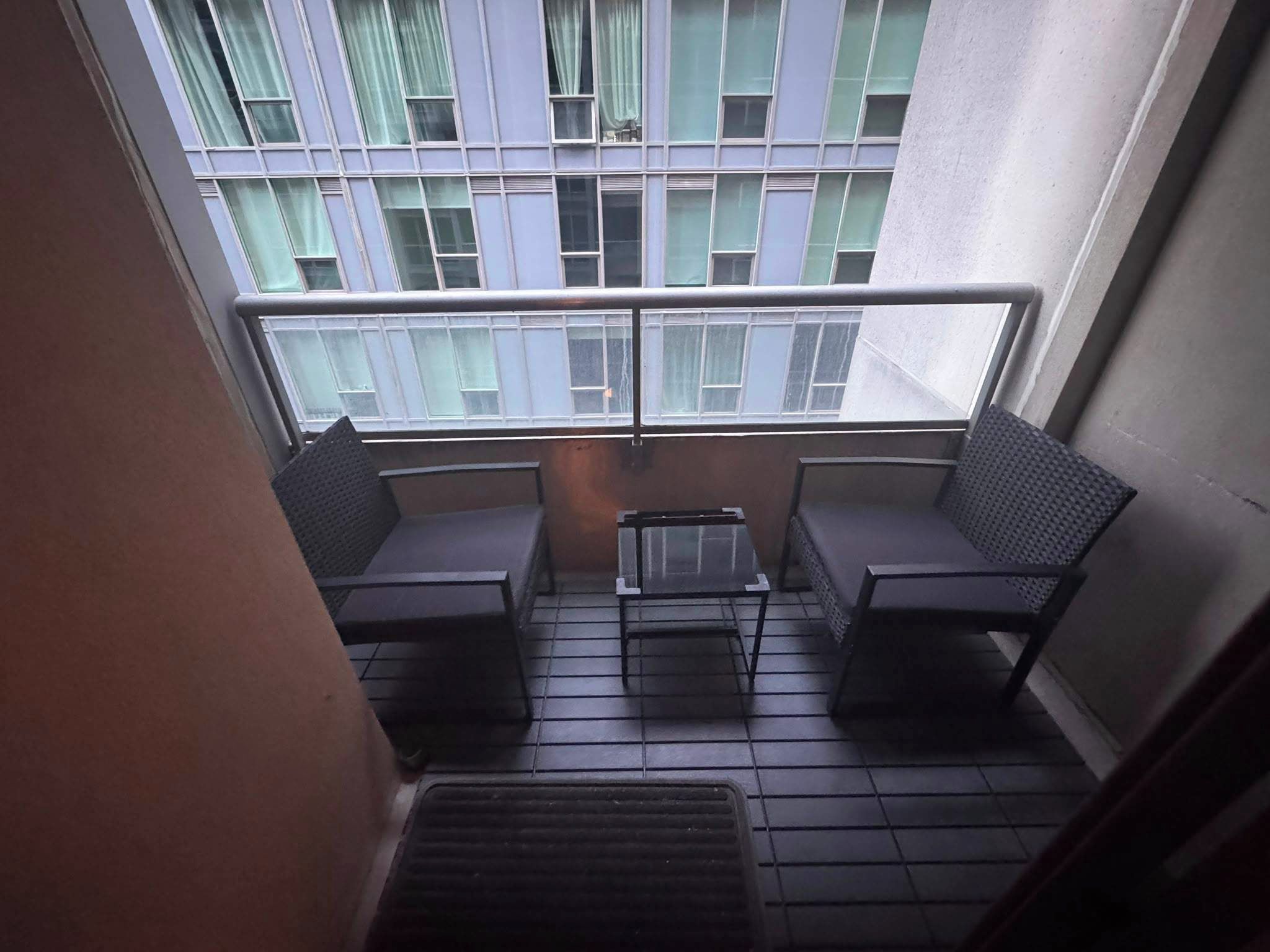 A small balcony with two chairs and a table, a window with curtains, and a metal grate on the floor.