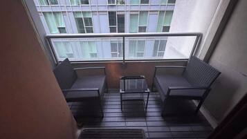 A small balcony with two chairs and a table, a window with curtains, and a metal grate on the floor.