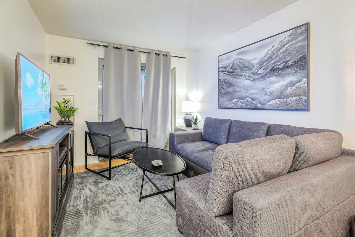 A modern living room with a grey sofa, a black armchair, a coffee table, and a flat-screen TV on a wooden cabinet.