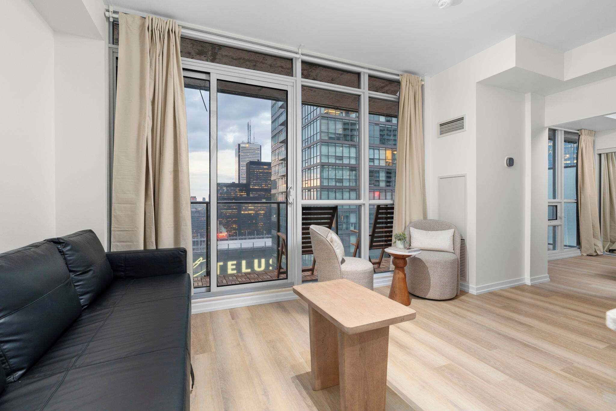 A modern living room with a black sofa, a wooden coffee table, and a chair. Large windows offer a city view.