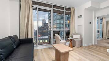 A modern living room with a black sofa, a wooden coffee table, and a chair. Large windows offer a city view.