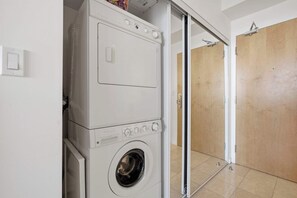A compact laundry area with a front-loading washing machine and dryer, a wooden door, and a tiled floor. - CN Tower View Condo -  Balcony, WiFi & Workspace (Toronto)