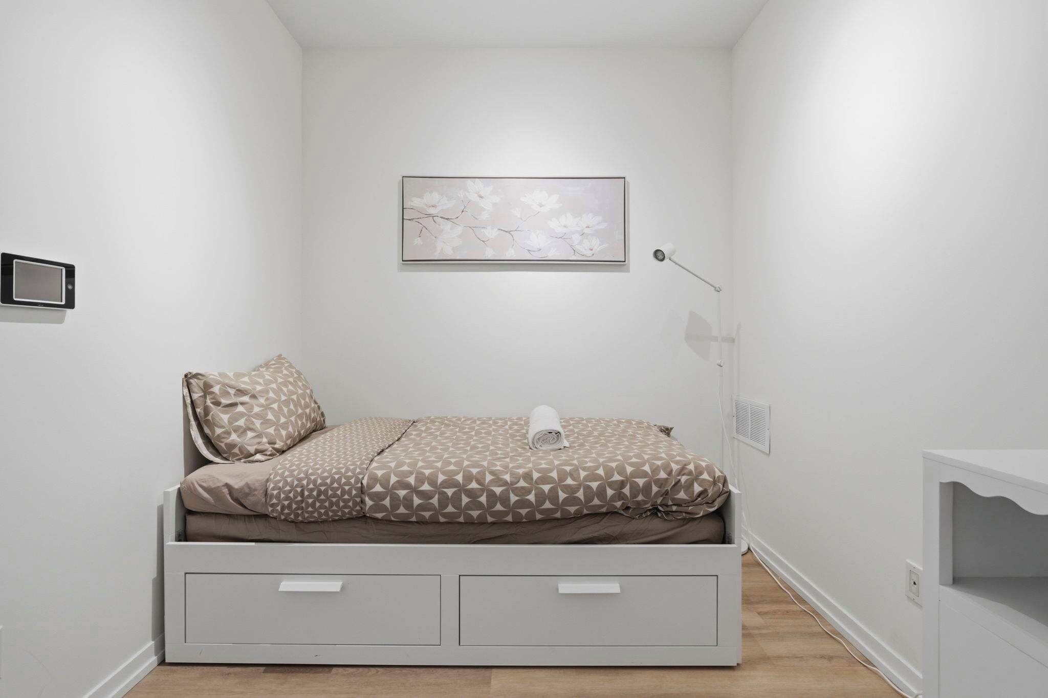 A minimalist bedroom with a bed, a wall-mounted artwork, a bedside lamp, and a small storage unit.