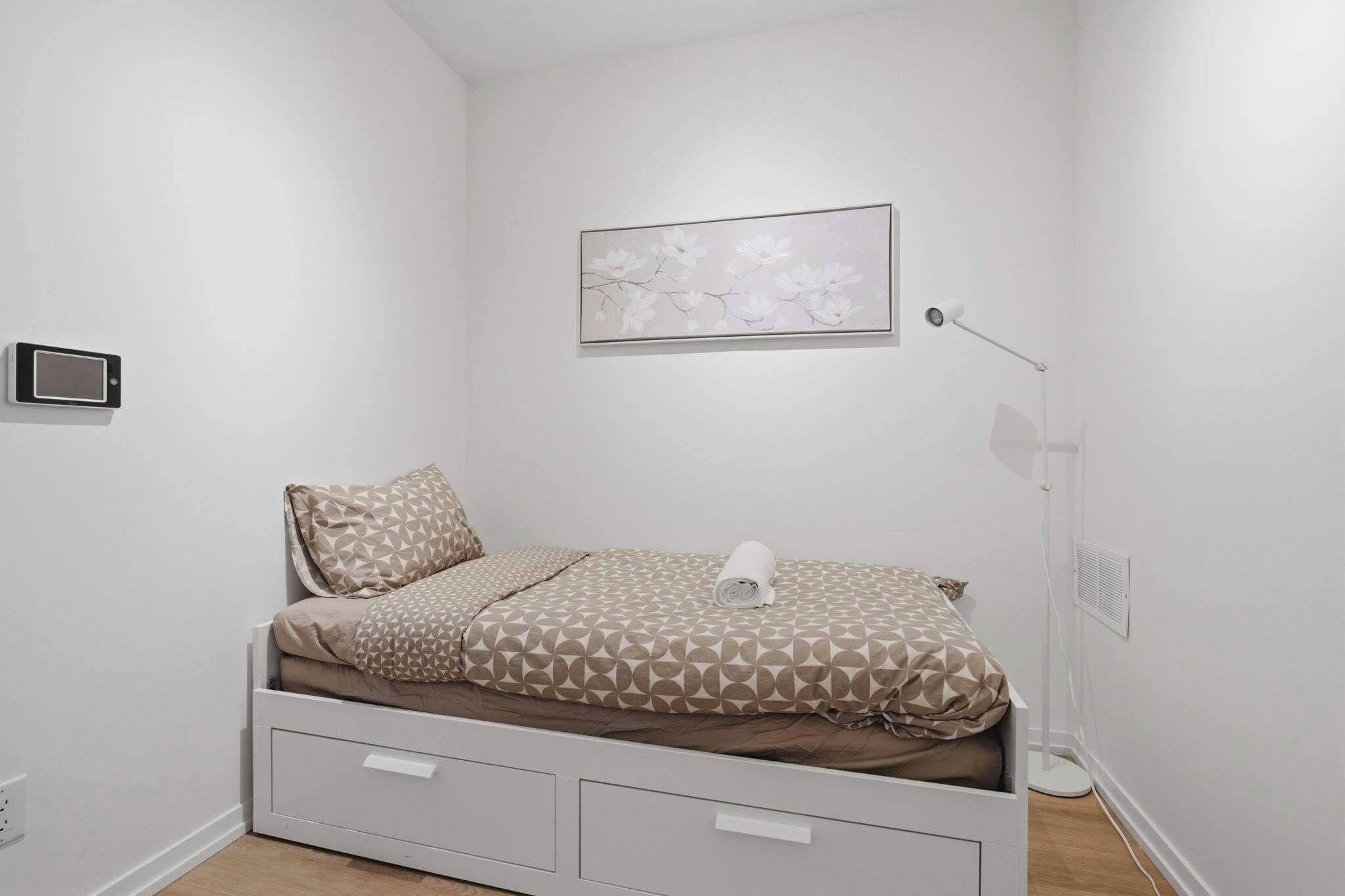 A small, neatly arranged bedroom with a bed, a wall-mounted picture, a bedside lamp, and a wall-mounted device.