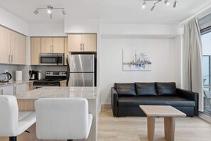 A modern kitchen with a stainless steel refrigerator, microwave, and stove. There is a black leather sofa and a small wooden table in the living area. - CN Tower View Condo -  Balcony, WiFi & Workspace (Toronto)