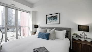 A modern hotel room with a large bed, a balcony with chairs, and a city view.