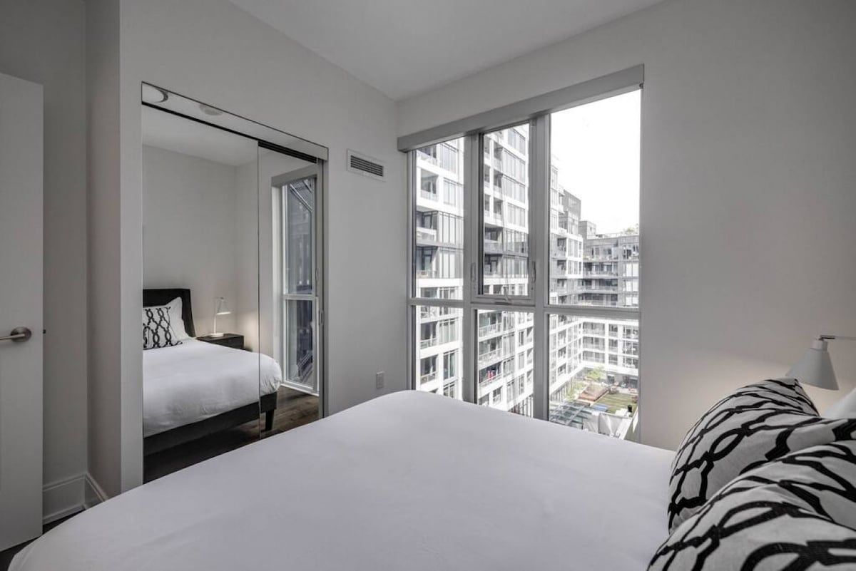 A hotel room with a large bed, a view of the city through floor-to-ceiling windows, and a door leading to another room.