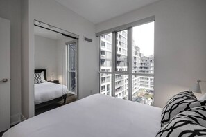 A hotel room with a large bed, a view of the city through floor-to-ceiling windows, and a door leading to another room.