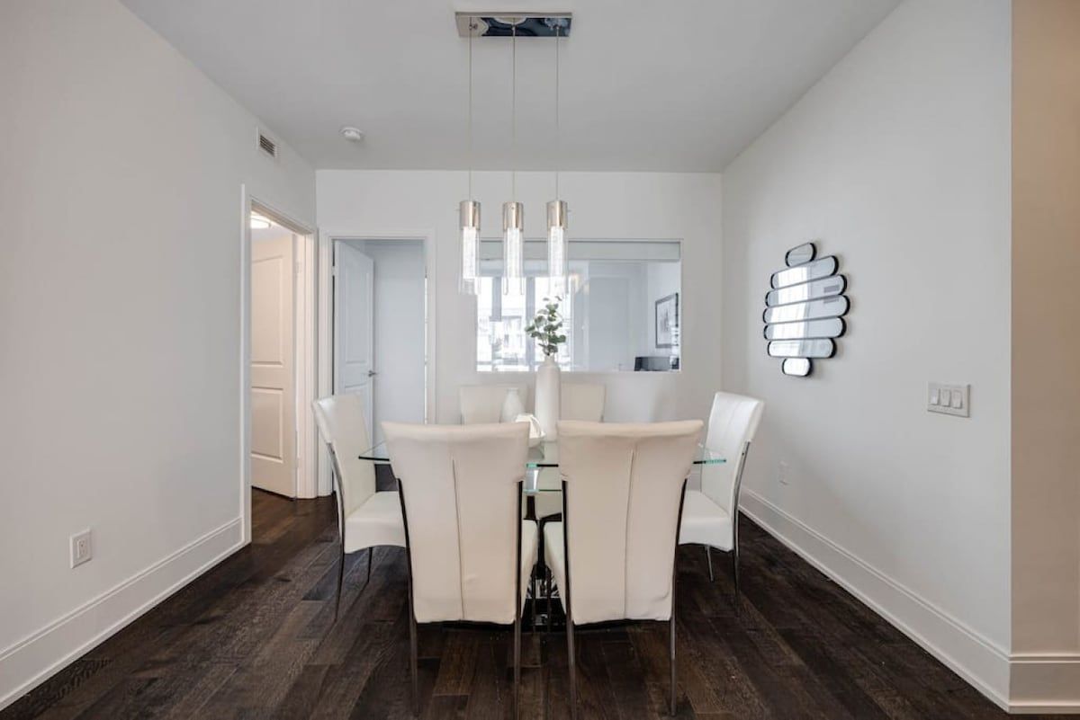 A dining area with a glass table and white chairs, a modern pendant light, and a wall-mounted light fixture.
