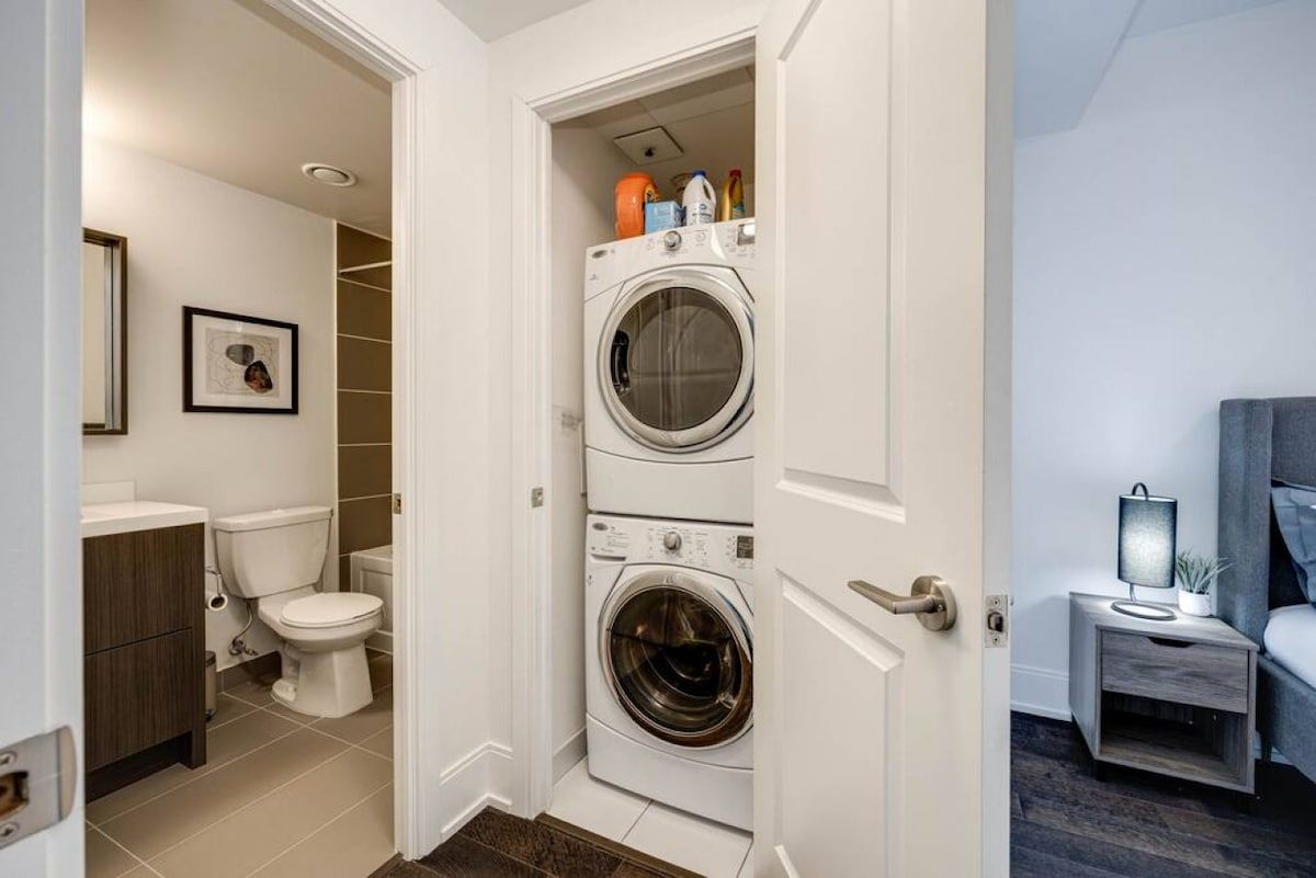 A laundry room with two stacked washing machines, a toilet, and a small bedside table with a lamp.