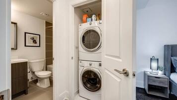 A laundry room with two stacked washing machines, a toilet, and a small bedside table with a lamp.