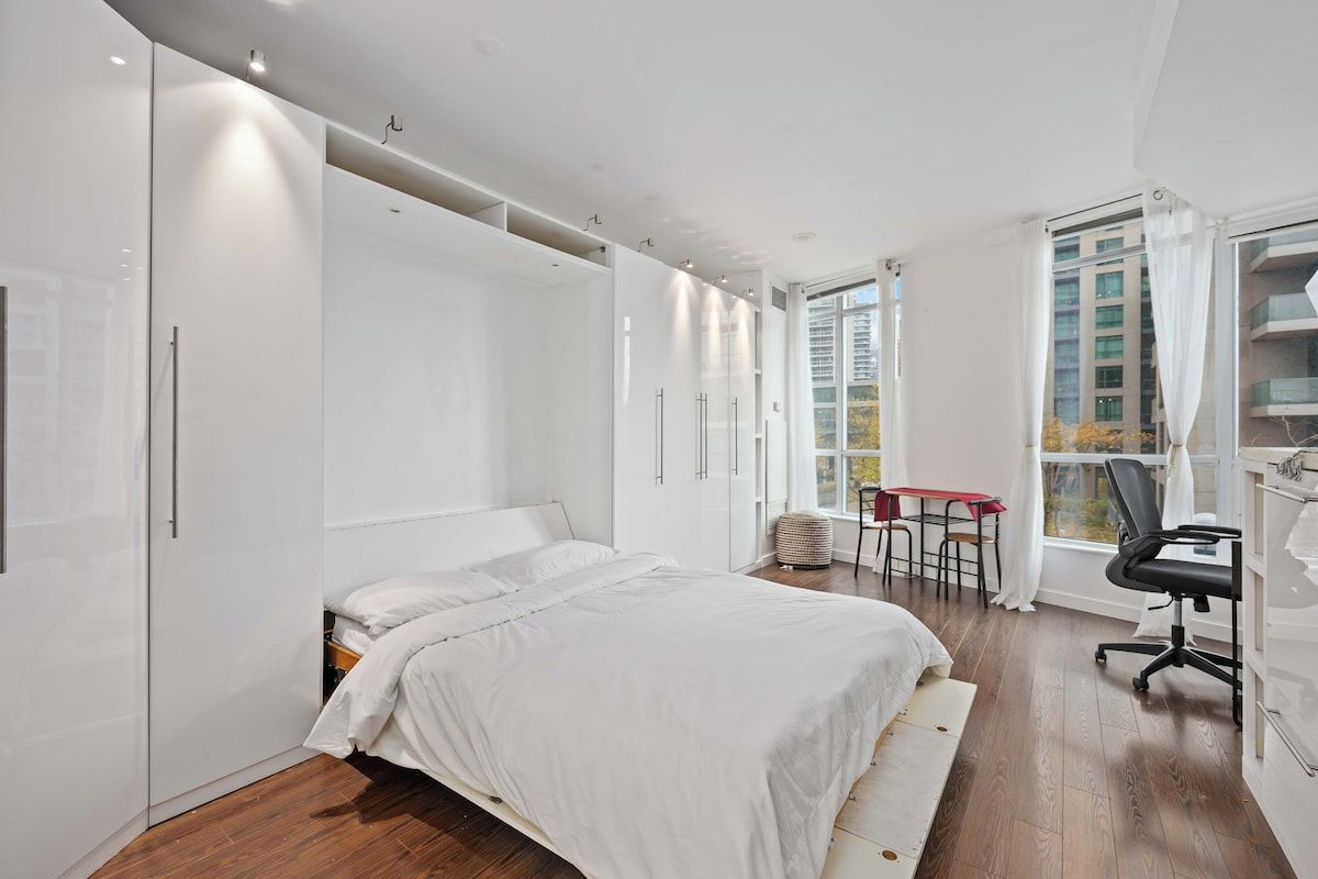 A bedroom with a bed, a desk, and a chair, with a window and a view of buildings outside.