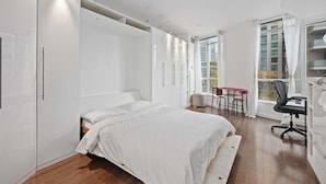 A bedroom with a bed, a desk, and a chair, with a window and a view of buildings outside. - Stylish Waterfront Studio Near Parks & City Core (Toronto)