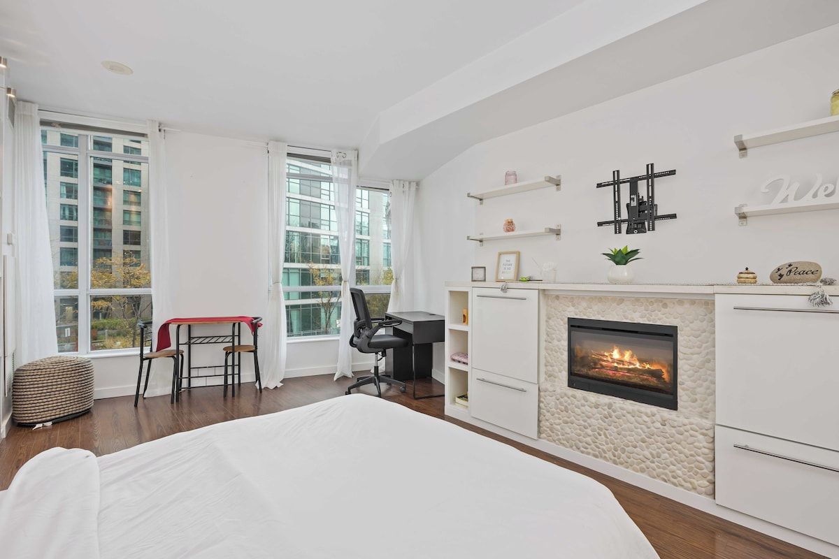 A bedroom with a bed, a desk, a chair, a fireplace, and large windows.
