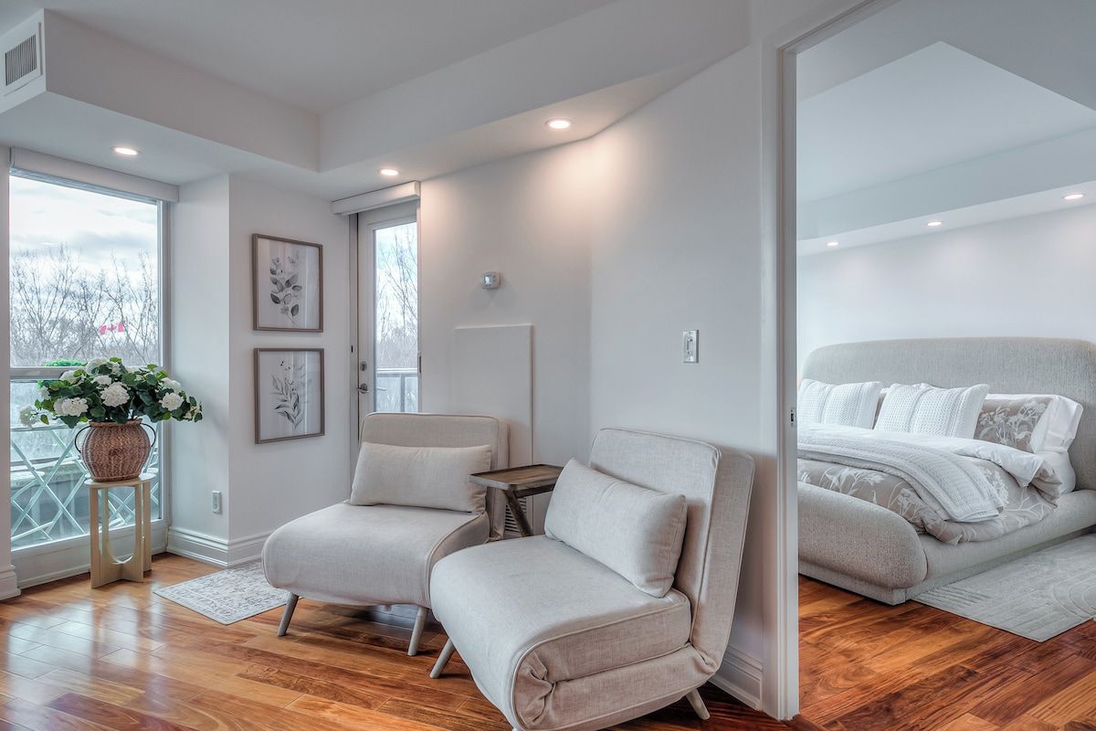 A modern bedroom with a large bed, two armchairs, a small table, and a window with a view of trees.