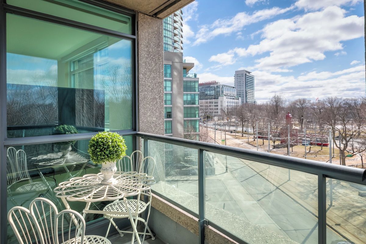 A balcony with a glass table and chairs, a potted plant, and a view of a cityscape with buildings and trees.