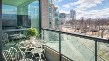 A balcony with a glass table and chairs, a potted plant, and a view of a cityscape with buildings and trees.