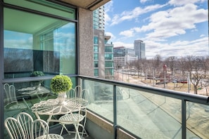 Un balcone con un tavolo e sedie in vetro, una pianta in vaso e vista su una città con edifici e alberi.